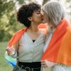 Two women hug under a Pride flag outdoors, smiling in celebration.