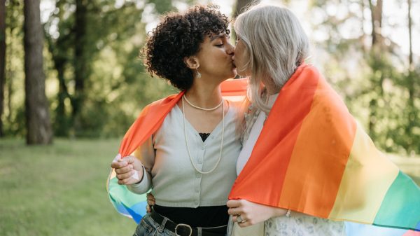 Two women hug under a Pride flag outdoors, smiling in celebration.