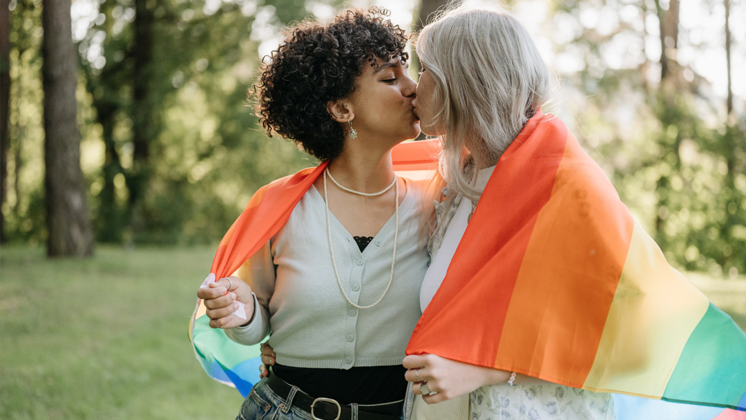 Two women hug under a Pride flag outdoors, smiling in celebration.